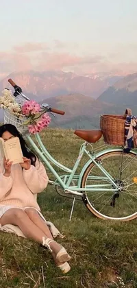 Get enchanted by this phone live wallpaper featuring a whimsical scene of a woman sitting on a grassy meadow with a bike, books, flowers, and picture frames beside her