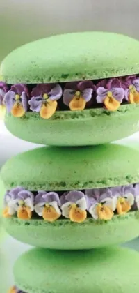 This lively phone wallpaper features three visually appealing green macarons stacked atop each other in a vibrant pastel hue, complemented by baroque-style violet flowers and delicately crafted fruit and floral designs