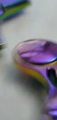 Looking for a mesmerizing live wallpaper for your phone? Check out this abstract fidget spinner design! Featuring a close up of a metallic spinner on a table, this wallpaper showcases random metallic colors in shades of purple and blue