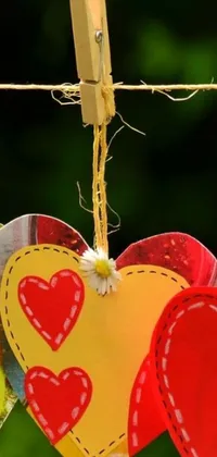 This phone live wallpaper features a charming design of heart-shape paper cutouts in a vibrant yellow and red color scheme, hanging on a clothes line amid a colorful, floral field