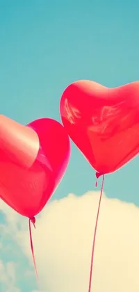 This live wallpaper features two heart-shaped balloons in brilliant red, floating gently in a clear blue sky amidst soft white clouds