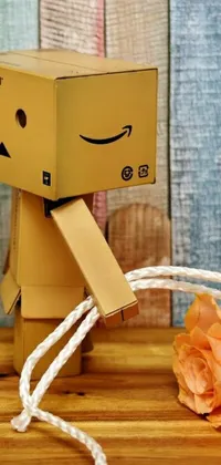 Get this creative live wallpaper for your phone today! Watch as a simple brown cardboard box sits atop a wooden table, while a neo-dada robot with a fishing rod and a cute smile stands by