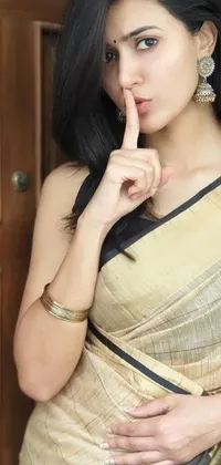 This stunning live phone wallpaper features a woman wearing a beige sari, with her finger placed delicately over her lips, as though keeping a secret