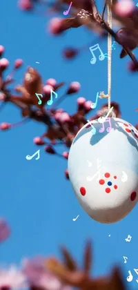 This decorative phone live wallpaper showcases a mesmerizing ornament hanging from a tree branch, embellished with musical notes that move softly in the wind
