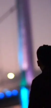 Looking for a visually thrilling phone live wallpaper? Look no further than this image of a man standing on a bridge at night, surrounded by a colorful haze that seems to come alive