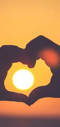 This phone live wallpaper captures a beautiful sunset scene with a person holding their hands in the shape of a heart