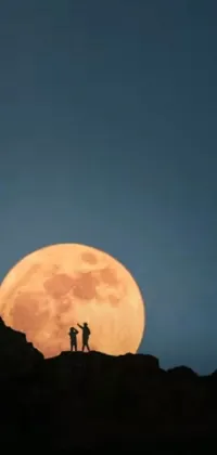 This live wallpaper depicts a serene and breath-taking view of two individuals standing atop a mountain under a bright and beautiful full moon