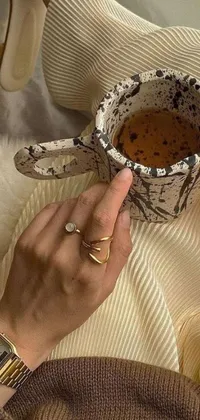 This phone live wallpaper features a close-up of a hand holding a hot cup of coffee adorned with mineral and gold jewelry