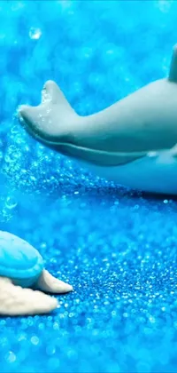 This vivid phone live wallpaper boasts a macro photograph of a dolphin and turtle figurine against a mesmerizing blue sand background