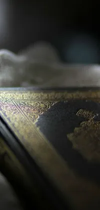 This phone live wallpaper showcases an old book with intricate Hurufiyya designs carved on its cover, placed on a wooden table
