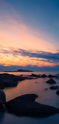 This live wallpaper depicts a serene sunset scene over water with rocks in the foreground