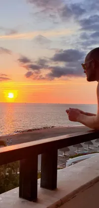 This stunning live wallpaper captures a beautiful sunset over the ocean, with a man standing thoughtfully by the water