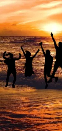 This stunning phone live wallpaper captures a candid moment of people jumping in the air at the beach as the evening sun sets behind them in a vibrant display of orange and yellow