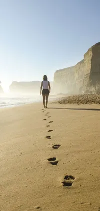 This phone live wallpaper features a stunning beach scene with footprints in the sand and a woman walking towards a cliff