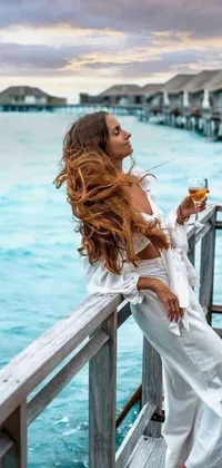 The phone wallpaper showcases a serene and inviting scene of a woman standing on a pier with a glass of wine