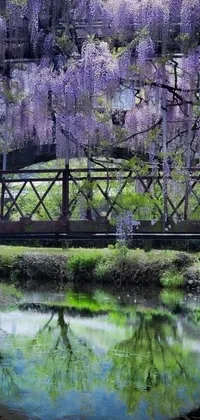 This live phone wallpaper is a stunning shot of a bridge over a body of water next to a lush green forest with purple flower trees
