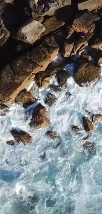 Experience the thrill of riding on waves with this surf-themed phone live wallpaper! Featuring an aerial view of a surfer on a rocky beach caught in action, the water's movement and waves crashing to the rocks are captured perfectly through the cinestill 800t 18mm lens
