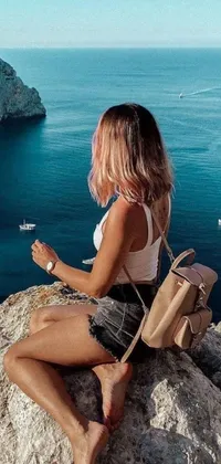 This phone live wallpaper depicts a young woman sitting atop a rocky ledge by the ocean with a backpack