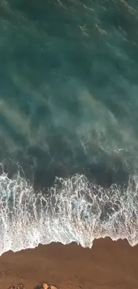 This phone live wallpaper features aerial footage of a sandy beach and ocean in a top-down perspective with waves, swirling particles, and a teal aesthetic