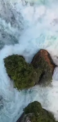 Decorate your phone background with a mesmerizing heart-shaped rock standing firmly in a river and surrounded by extremely humongous waves that relentlessly crash onto it