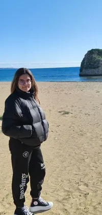 A woman stands atop a sandy beach, wearing jeans and a black hoodie with a backpack and camera slung over her shoulder in this phone live wallpaper
