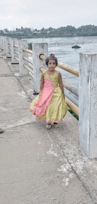 This phone live wallpaper depicts an adorable little girl in a vibrant pink and yellow dress, standing on a bridge against a serene landscape