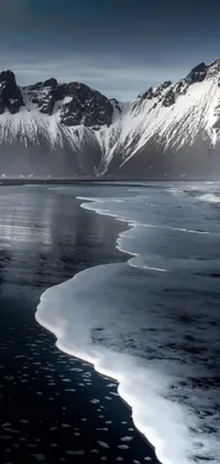 This phone live wallpaper showcases a serene black sand beach with snow-capped mountains in the backdrop