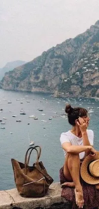 This phone live wallpaper showcases a stunning image of a woman sitting atop a stone wall near the ocean