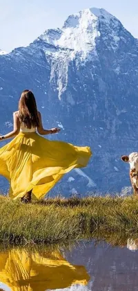 This phone wallpaper shows a woman in a yellow dress next to a cow in a scenic field