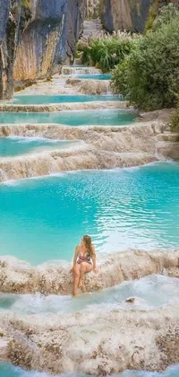 This phone live wallpaper showcases a beautiful woman sitting on the edge of a stunning water pool, surrounded by a picturesque landscape