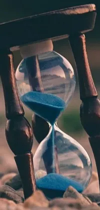 This phone live wallpaper features an hourglass placed on an assortment of rocks; its sand consistently in motion, with background unsplash process art and an abstract timeline nexus embodied in a 3D close-up of the hourglass against a wooden background