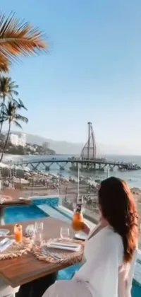 This phone live wallpaper displays a calming beach scene with a woman sitting at a wooden table beside a shimmering pool