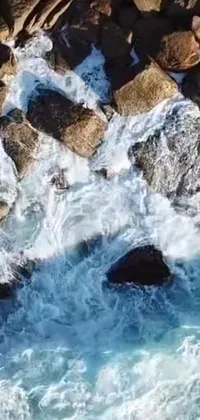 Get mesmerized by this phone live wallpaper that portrays a stunning bird's eye view of undulating waves in a vast blue body of water, bounded by rocks