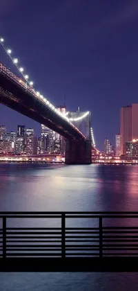 Experience the beauty of a bridge extending over calm waters with the stunning cityscape illuminated behind it through this live wallpaper