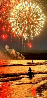 This phone live wallpaper showcases a picturesque beach scene, featuring a group of people standing by the ocean with a magnificent fireworks display lighting up the night sky behind them