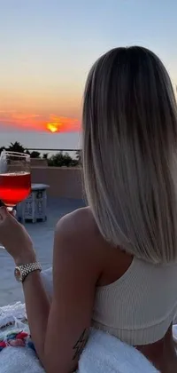 This stunning live wallpaper depicts a blonde woman enjoying a glass of wine at a table while appreciating a picturesque image in the background