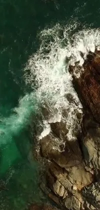 This phone live wallpaper features a stunning aerial shot of a large rock being hit by waves in the middle of a body of water