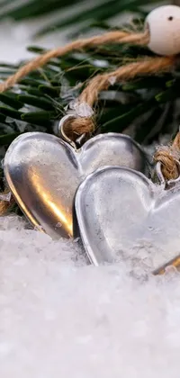 Get in the winter romance mood with this stunning phone live wallpaper! Featuring two silver hearts placed on a snow-covered ground surrounded by evergreen branches and a snowy landscape in the background, this beautiful and peaceful image is perfect for your phone screen